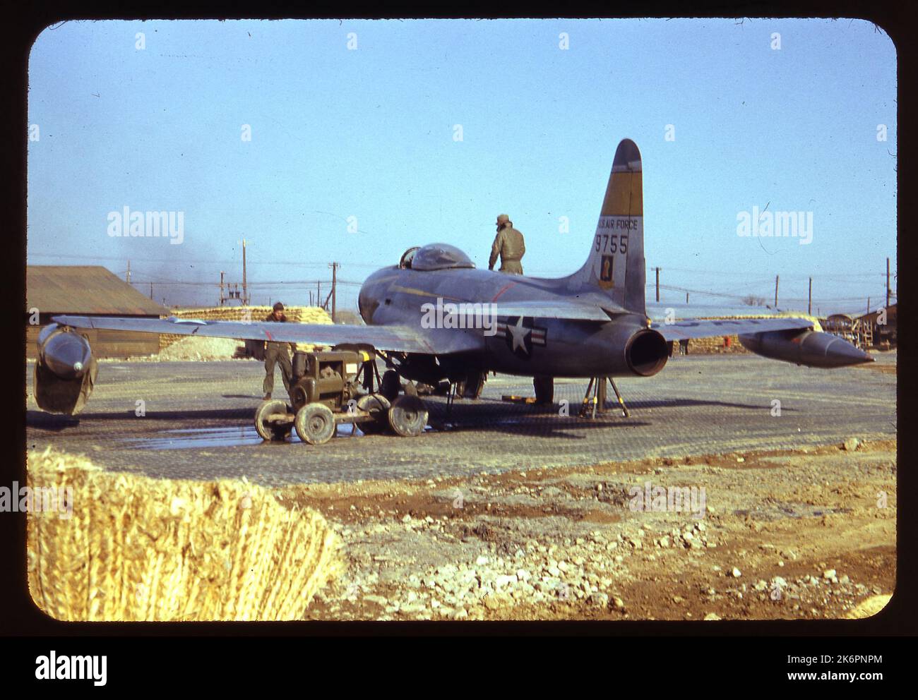 Vue arrière à la moitié gauche d'une étoile de tir Lockheed F-80C (n° de série 49-755) portant les marquages du 41st Fighter Interceptor Squadron (FIS), 35th Fighter Interceptor Wing (FIW), quelque part en Corée. L'avion est stationné devant des sacs de sable et un bâtiment. Un homme d'équipage se tient sur l'aile droite et un autre au sol sur le côté droit de l'avion. Un troisième homme se tient devant l'avion. Un véhicule de support est stationné sur le côté gauche et l'équipement est dispersé au sol. En arrière-plan, de la fumée est visible sur plusieurs bâtiments, des tas de sacs de sable et de l'équipement. Banque D'Images