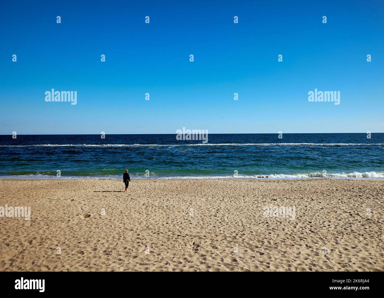 Immense plage, bleu mer et ciel et une jeune femme solitaire marchant le long de la plage vers la mer Banque D'Images