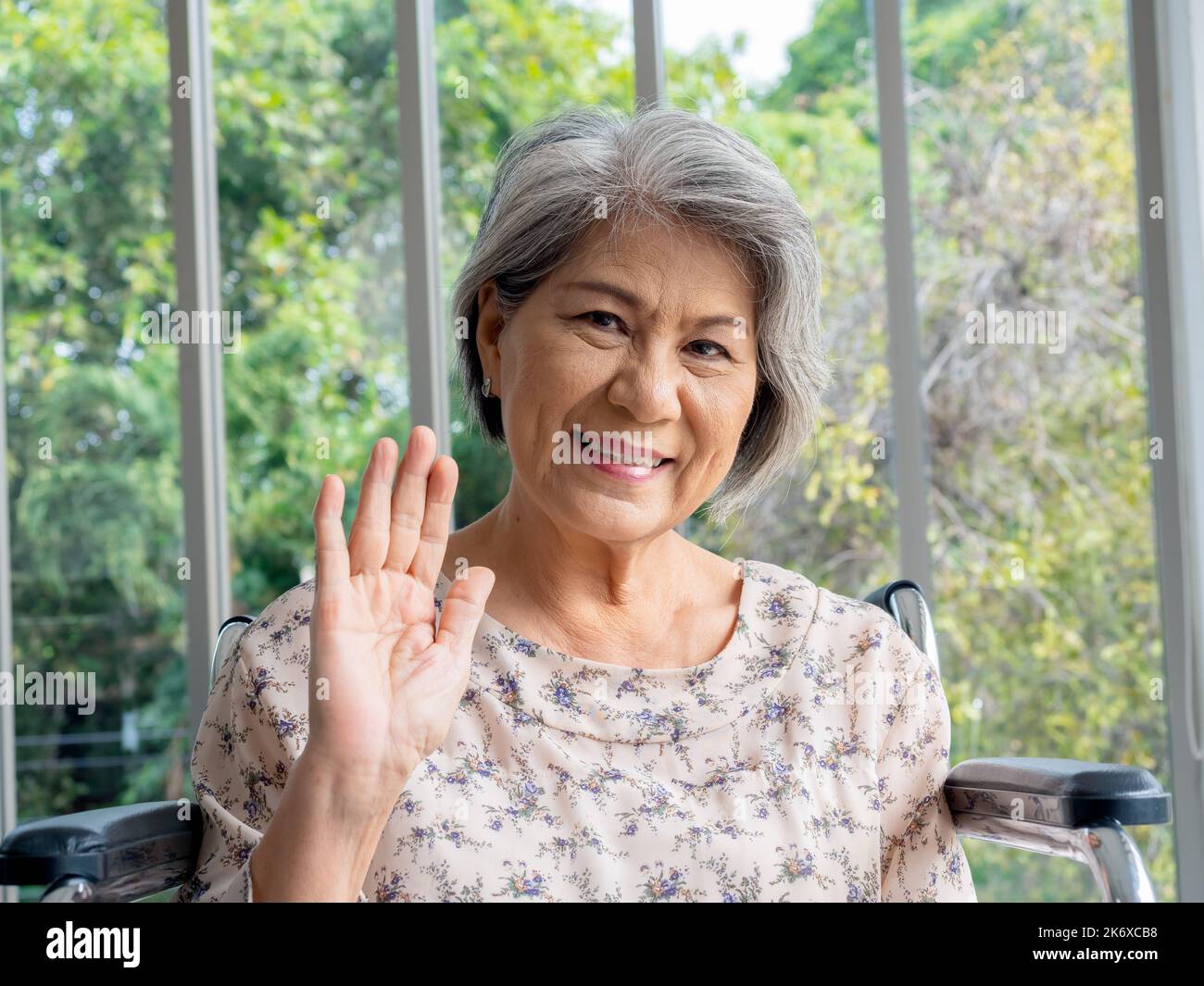 Portrait d'une femme asiatique âgée heureuse cheveux gris assis sur un fauteuil roulant, agitant salutation, souriant et regardant l'appareil photo. Une femme âgée rencontre sa famille Banque D'Images