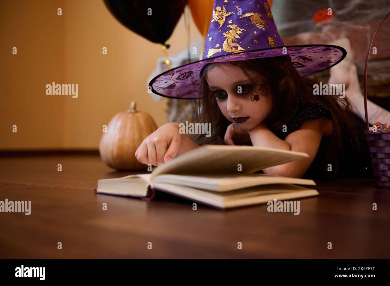 Petite fille espiègle avec maquillage Halloween vêtue de sorcière, sorcière dans le chapeau de magicien lisant un livre de sorcellerie Banque D'Images