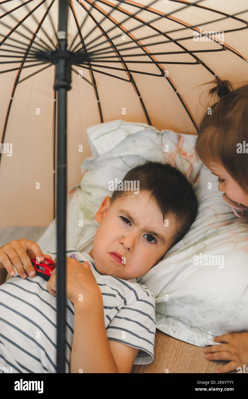 Garçon criant à la caméra tout en étant allongé dans la chambre sous un parapluie à côté de sa sœur. Fête d'enfants. Famille amusante. Banque D'Images