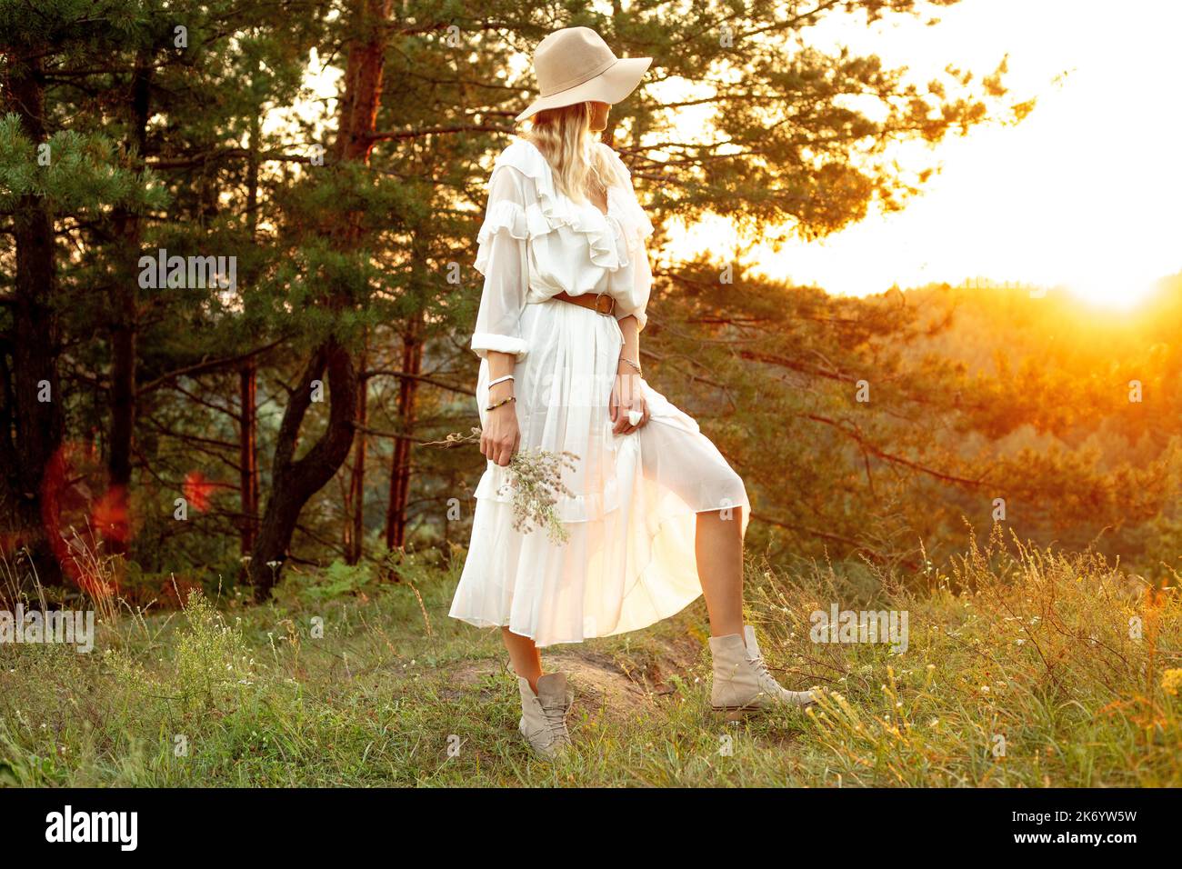 Femme blonde non reconnaissable en chapeau et robe tenant un bouquet d'herbes, se tenir sur la prairie près de la forêt contre le ciel de coucher de soleil Banque D'Images