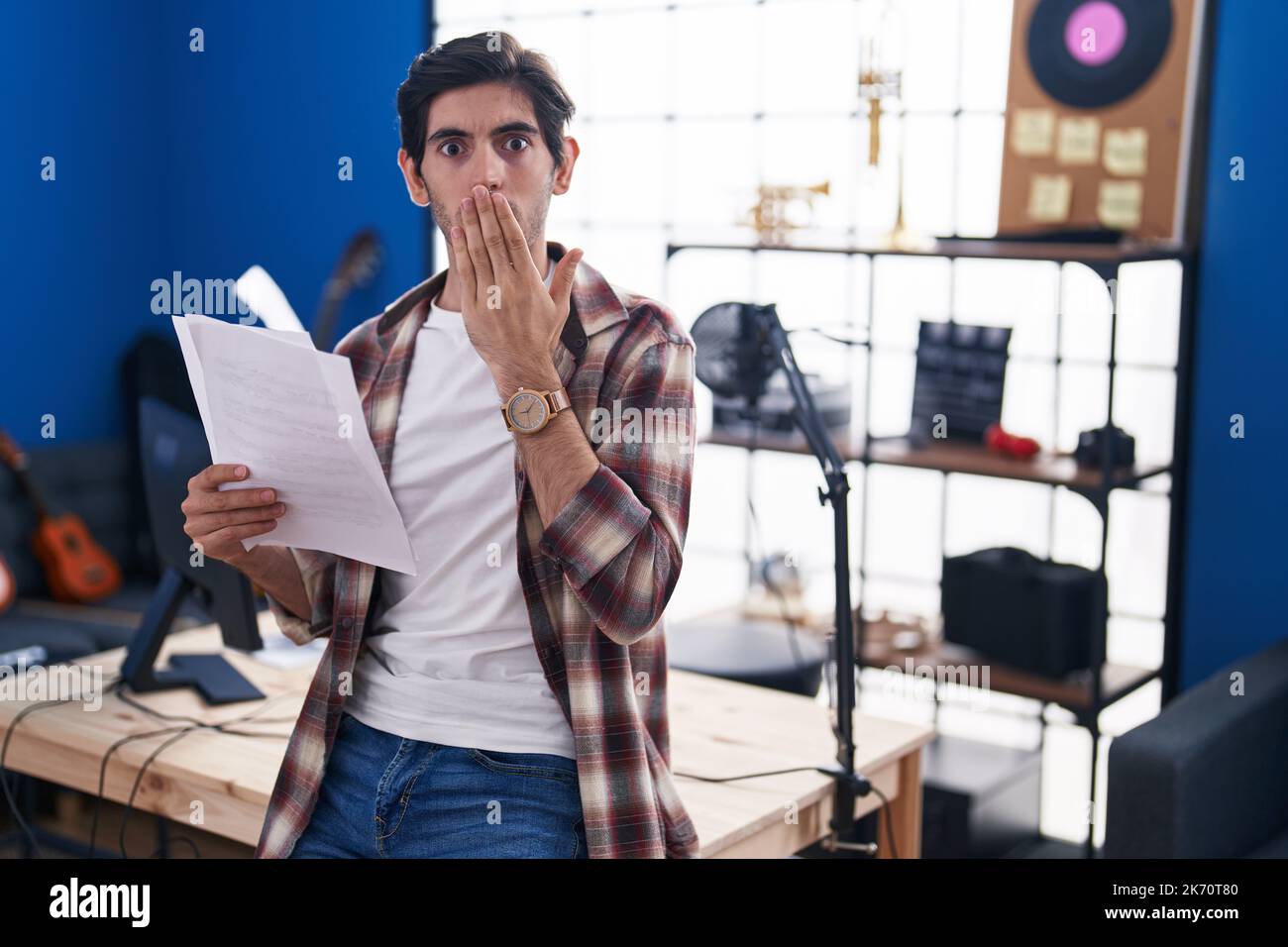 Jeune hispanique lisant la feuille de musique au studio de musique couvrant la bouche avec la main, choqué et peur de l'erreur. expression surprise Banque D'Images