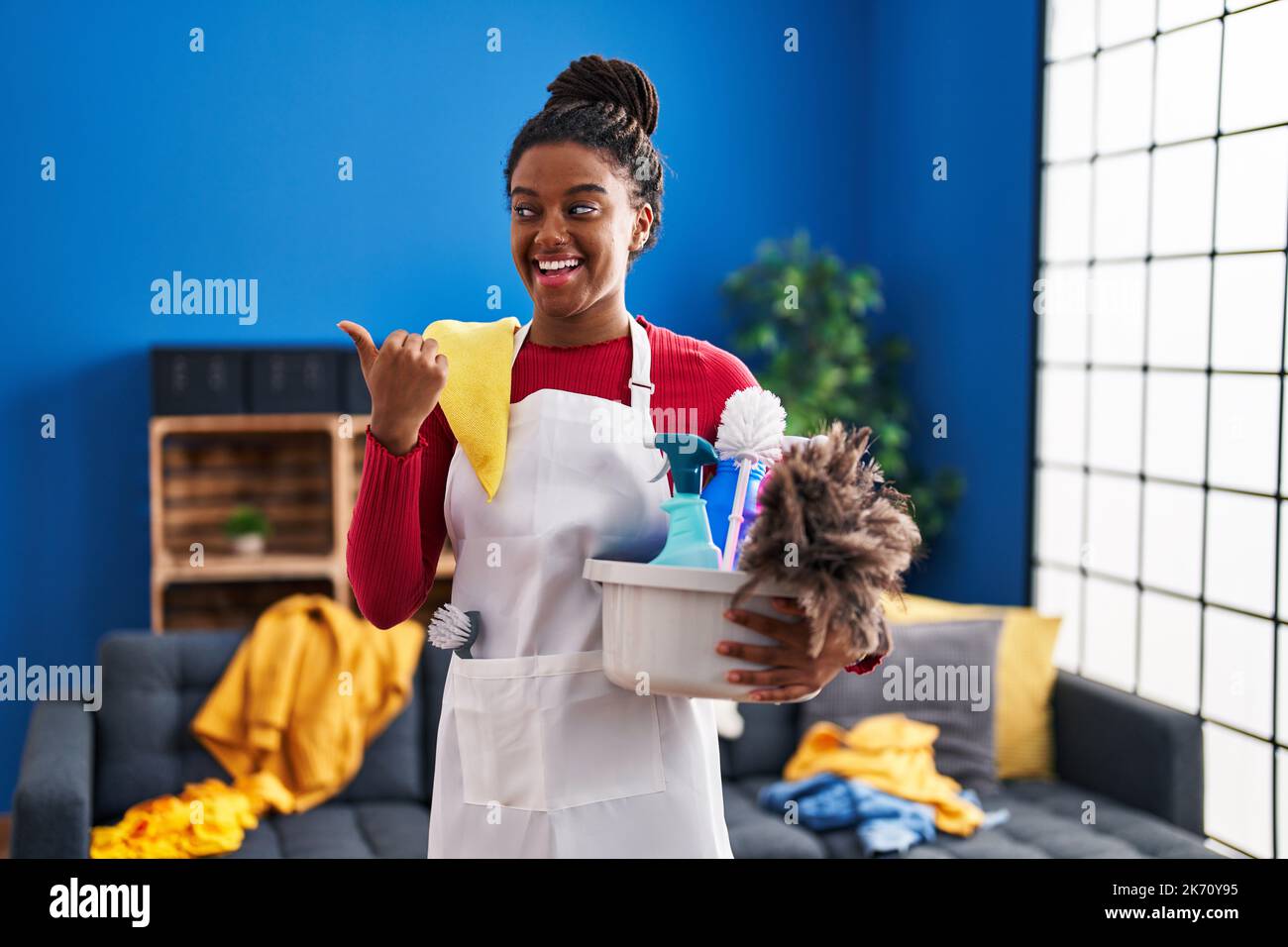 Jeune afro-américain avec des tresses de nettoyage de la maison tenant des produits de nettoyage pointant le pouce vers le côté souriant heureux avec la bouche ouverte Banque D'Images