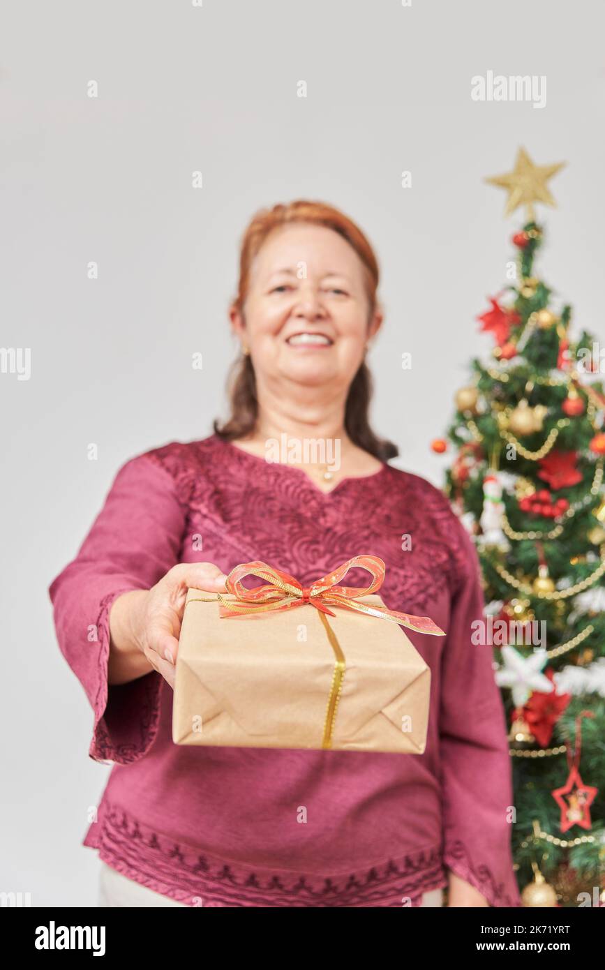 Femme hispanique mûre souriante alors qu'elle remet un cadeau de Noël à la caméra. Concept: La joie de donner des cadeaux pendant les vacances. Composition wi Banque D'Images