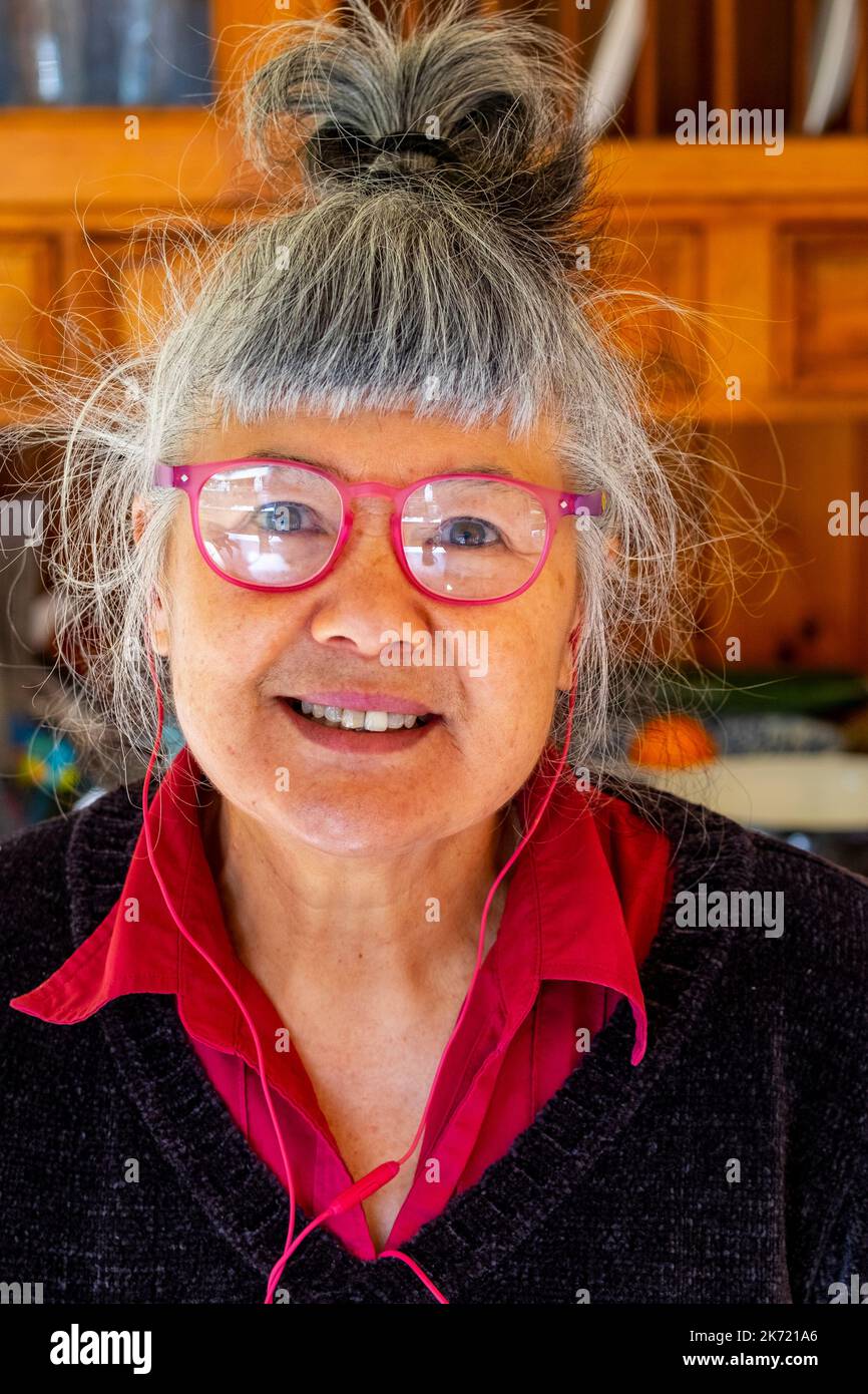 Femme de 68 ans d'origine chinoise/italienne avec lunettes de lecture rouges et écouteurs filaires Banque D'Images
