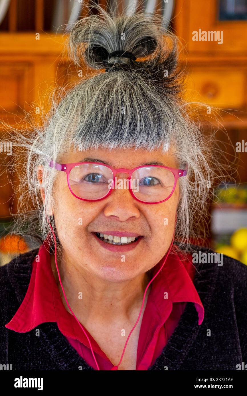 Femme de 68 ans d'origine chinoise/italienne avec lunettes de lecture rouges et écouteurs filaires Banque D'Images