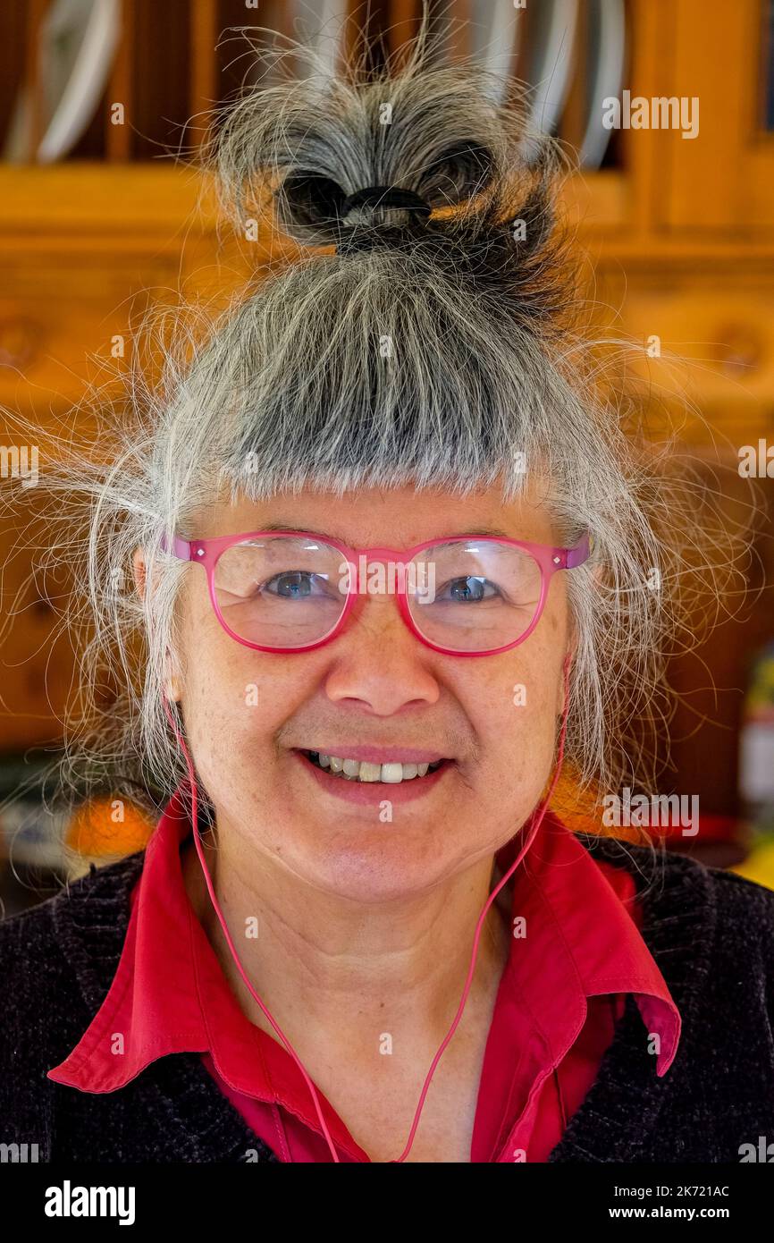Femme de 68 ans d'origine chinoise/italienne avec lunettes de lecture rouges et écouteurs filaires Banque D'Images