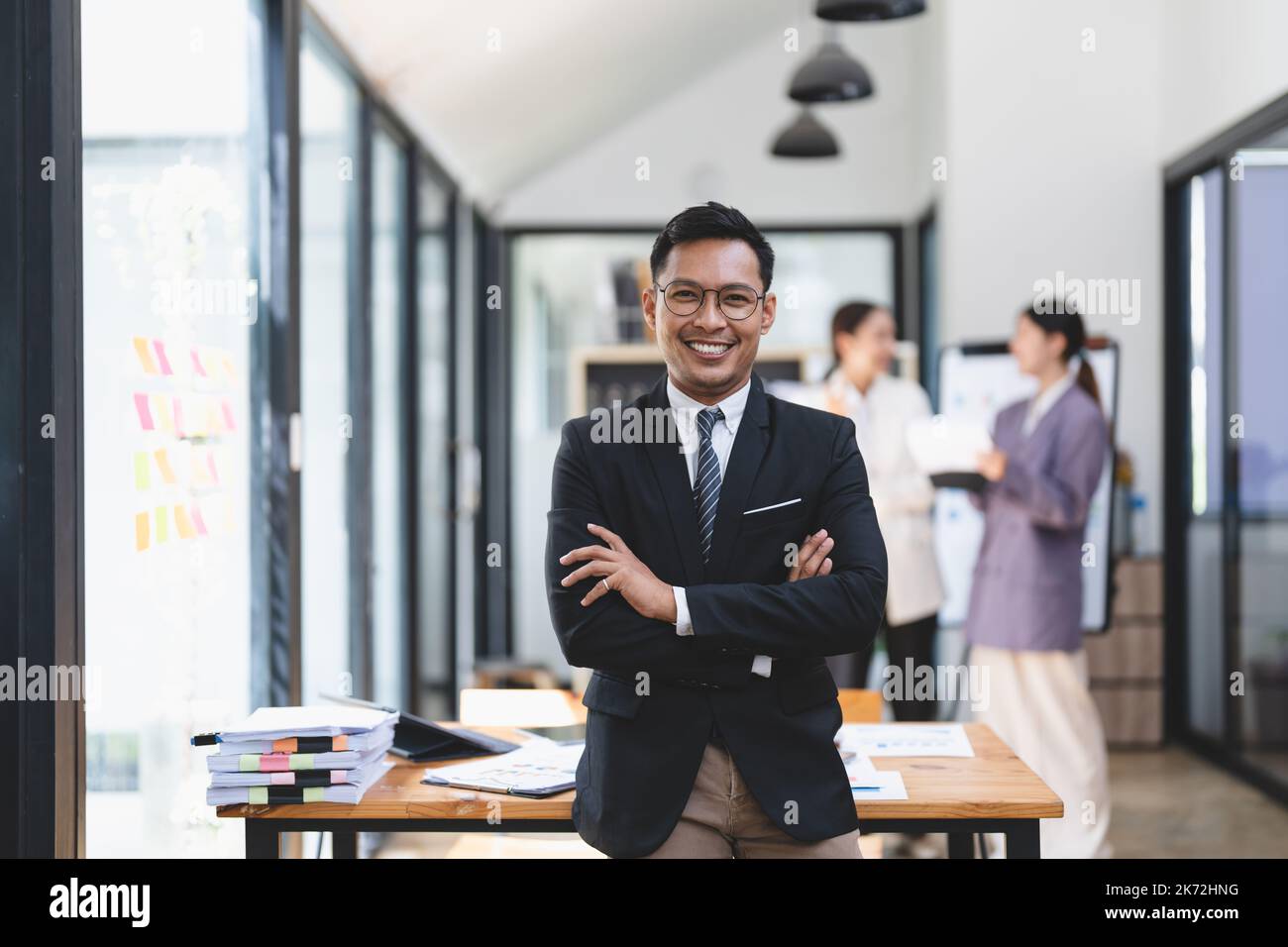 Portrait de la réussite asiatique homme de bureau dans les affaires formelles costumes sourire à l'appareil photo dans le bureau avec des collègues flous travaillant dans le bureau comme Banque D'Images