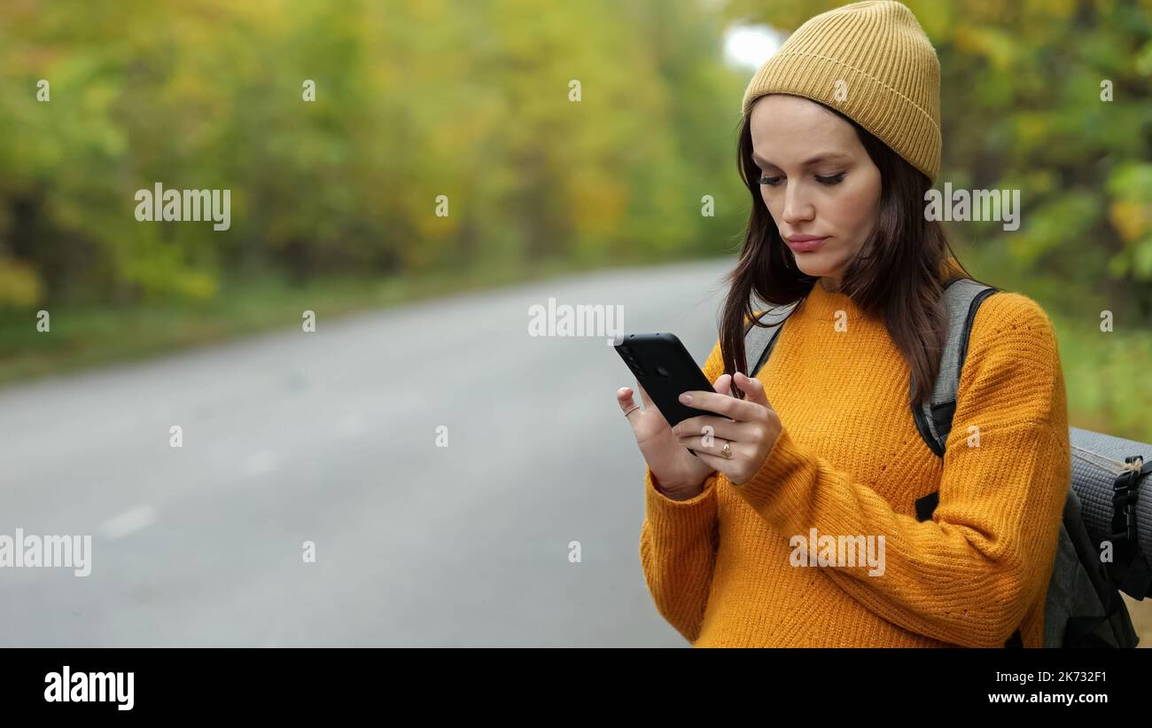 Une femme randonneur dans un chandail orange marche sur une route vide et saisit un message à un ami sur le téléphone. Banque D'Images