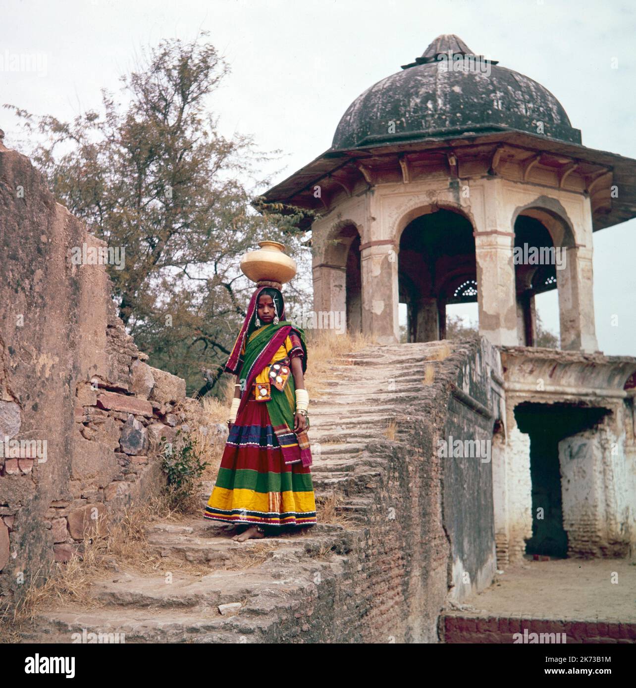 Vintage 1961 photo couleur montrant une jeune fille indienne qui vient de recueillir l'eau d'un puits et porte l'eau dans une casserole sur sa tête, en bas quelques pas. Banque D'Images