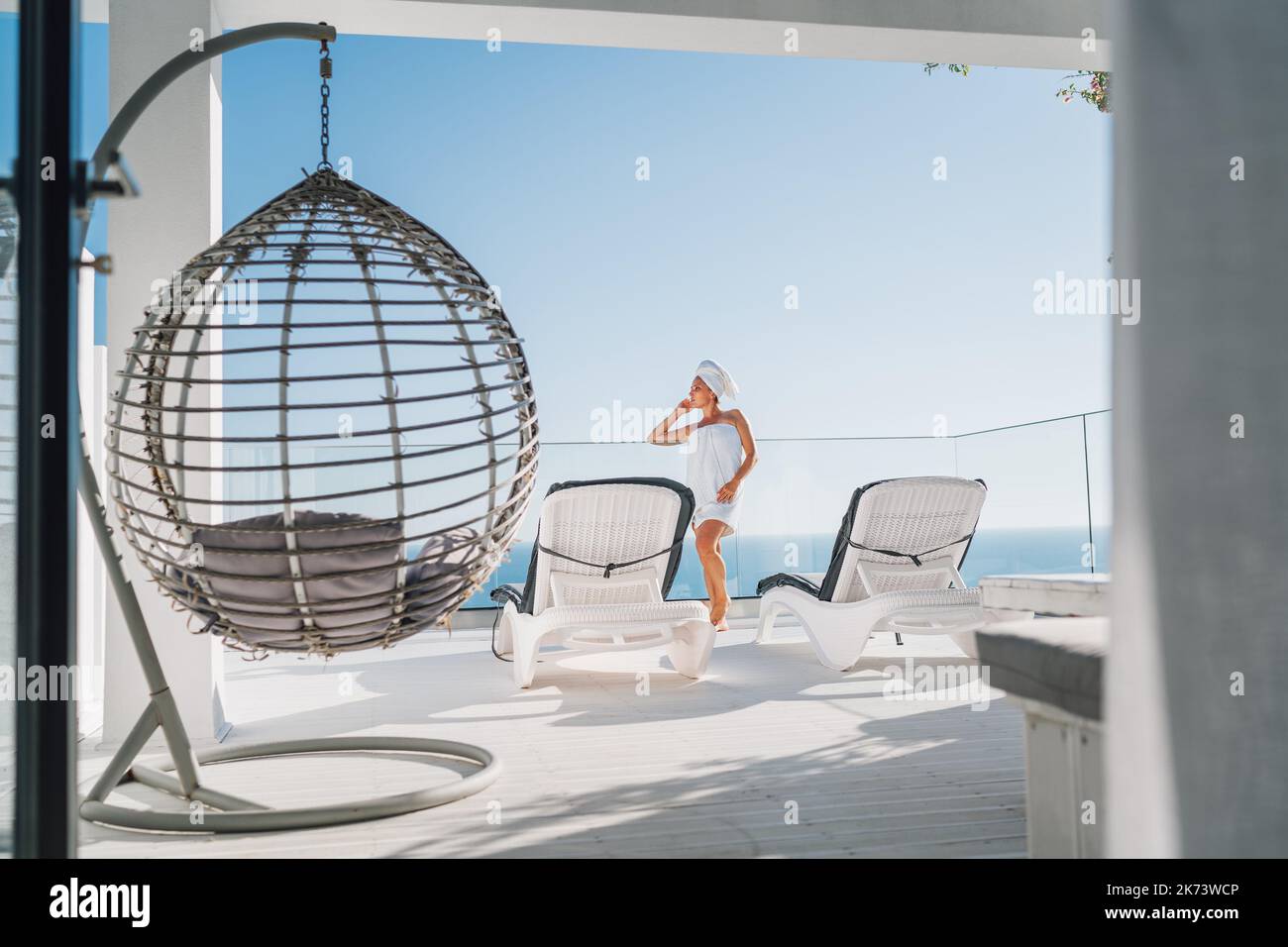 Après la douche belle femme avec cheveux et serviette de corps envelopper debout sur la terrasse de maison ouverte avec chaises longues, chaise suspendue appréciant paysage marin et bleu propre Banque D'Images