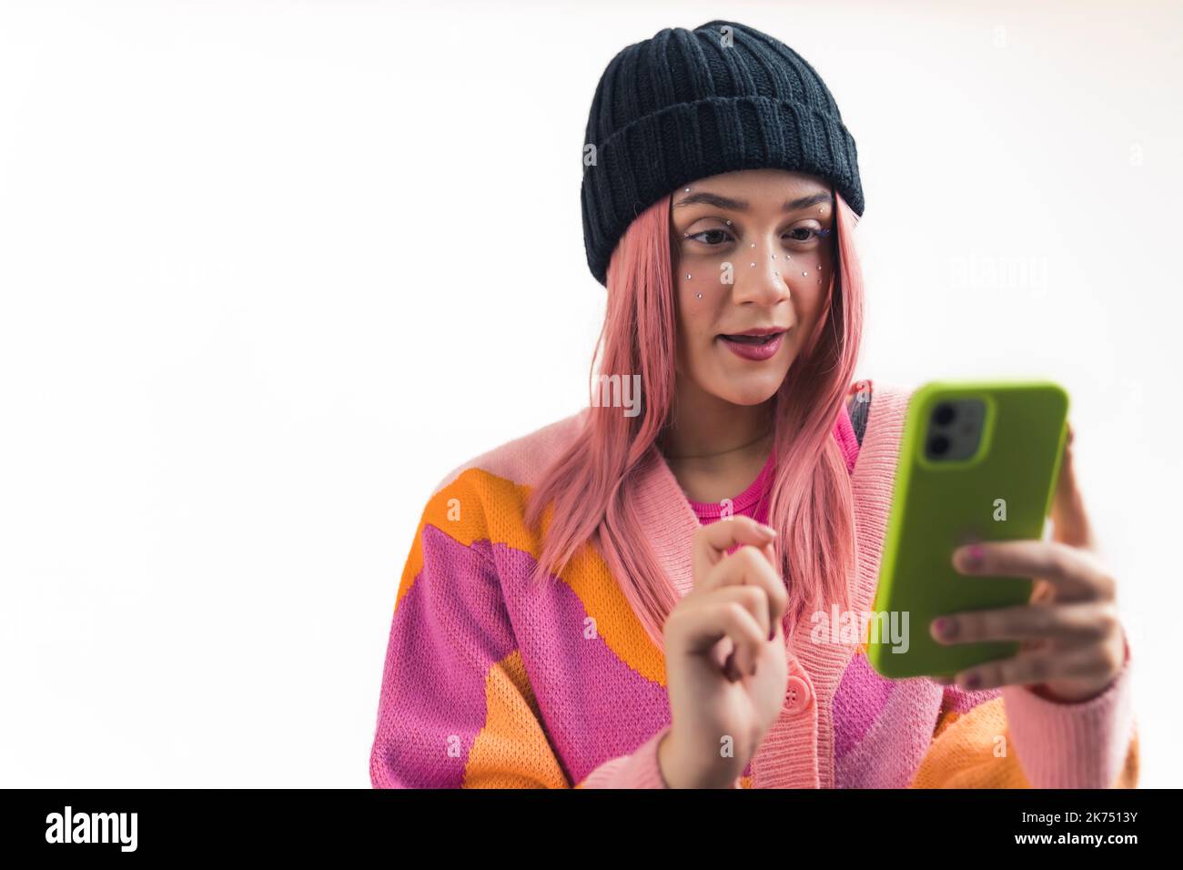 Jeune, joyeuse, élégante, caucasienne fille avec de longs cheveux roses, portant du maquillage, des vêtements colorés et un chapeau, tenant son téléphone devant son visage et regardant quelque chose sur l'Internet, isolé sur fond blanc. Photo de haute qualité Banque D'Images