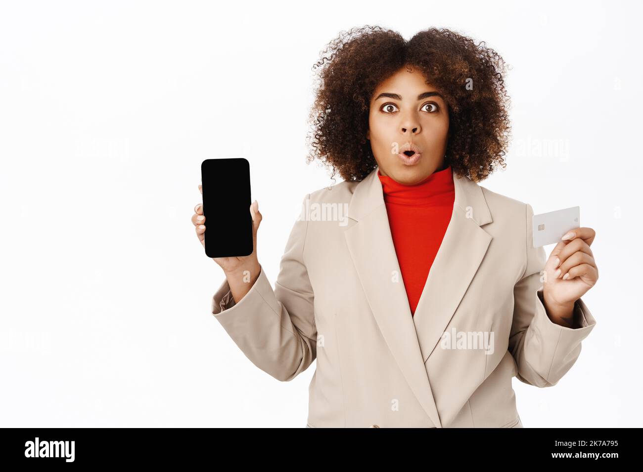 Une femme afro-américaine choquée montre son écran de téléphone mobile et sa carte de crédit, se tient en costume d'affaires, fond blanc Banque D'Images