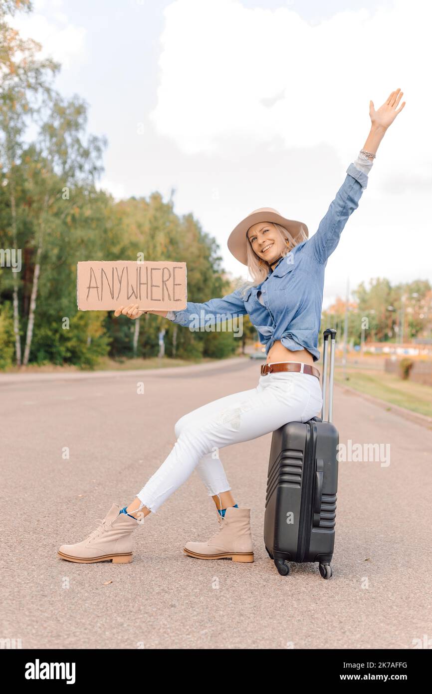 Une femme joyeuse essaie d'arrêter de passer une voiture assise sur une valise avec une affiche en carton étirée sur une autoroute vide. Dame en chapeau échapper de la ville à partir Banque D'Images