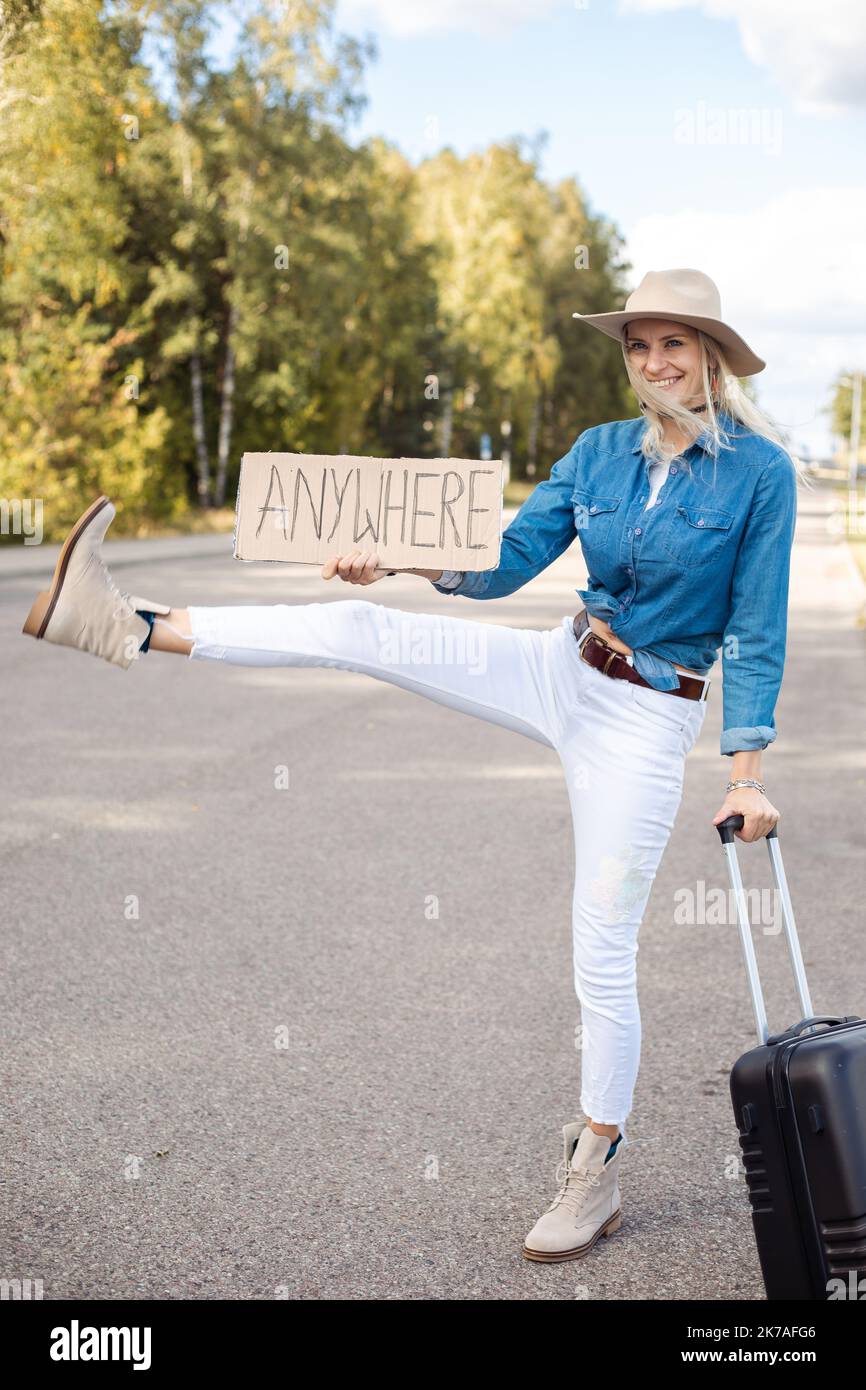 Joyeuse femme avec une valise essayer d'arrêter de passer la voiture avec une jambe tendue relevée et une affiche en carton sur une autoroute vide. Lady in Hat échapper de la ville à Banque D'Images