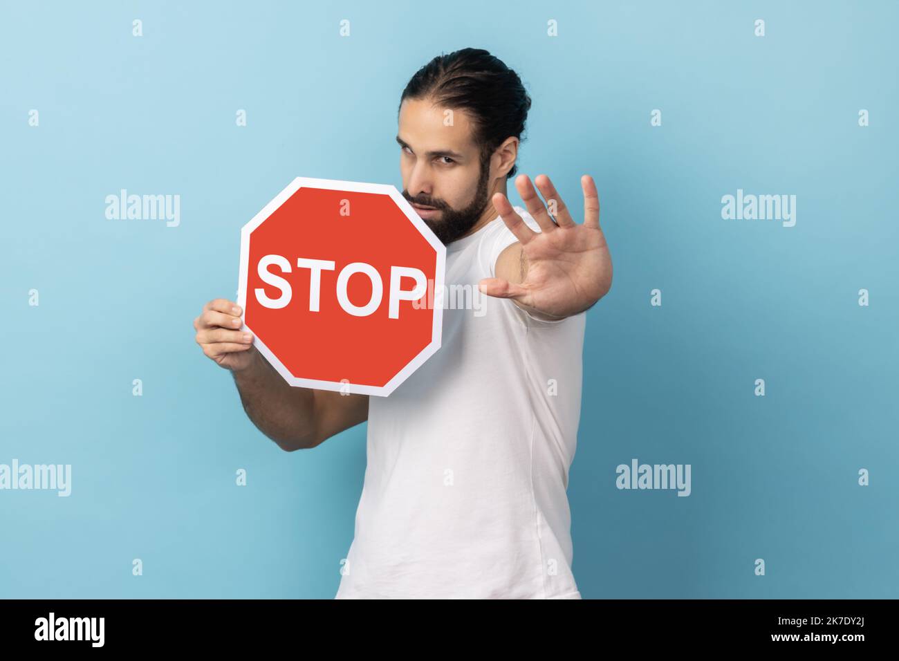 Portrait d'un homme strict avec une barbe portant un T-shirt blanc montrant un geste d'arrêt et tenant un panneau rouge d'arrêt, interdictions et restrictions. Studio d'intérieur isolé sur fond bleu. Banque D'Images