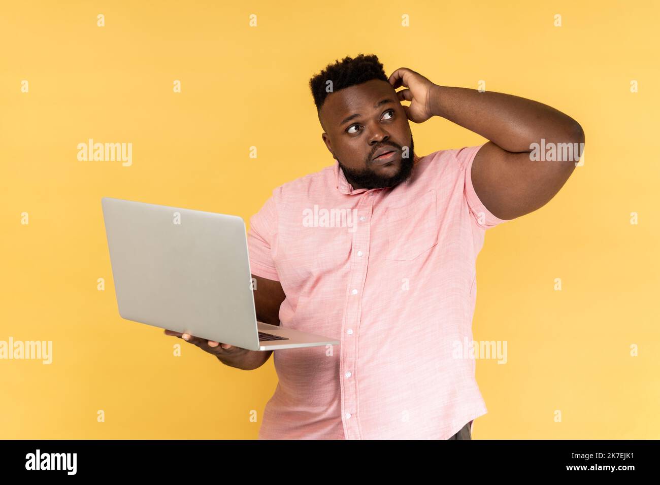 Portrait d'un homme barbu attentif portant une chemise rose tenant un ordinateur portable et pensant à la stratégie de démarrage avec une expression douteuse sérieuse. Studio d'intérieur isolé sur fond jaune. Banque D'Images
