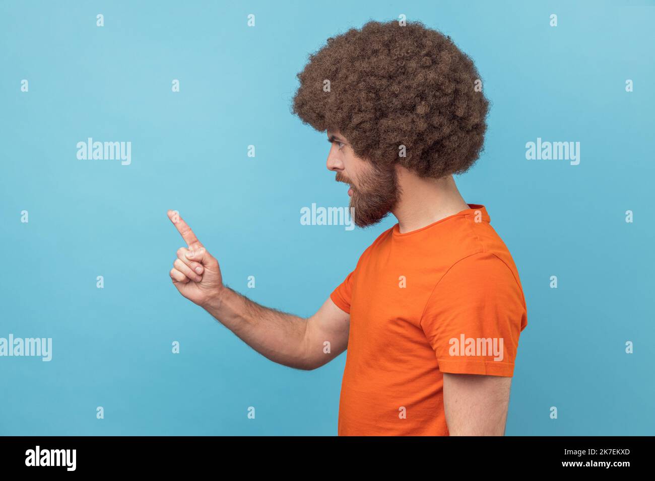 Vue latérale d'un homme sérieux avec une coiffure afro portant un T-shirt orange debout avec un geste d'admonishing, en faisant des signes d'erreur et d'avertissement. Studio d'intérieur isolé sur fond bleu. Banque D'Images