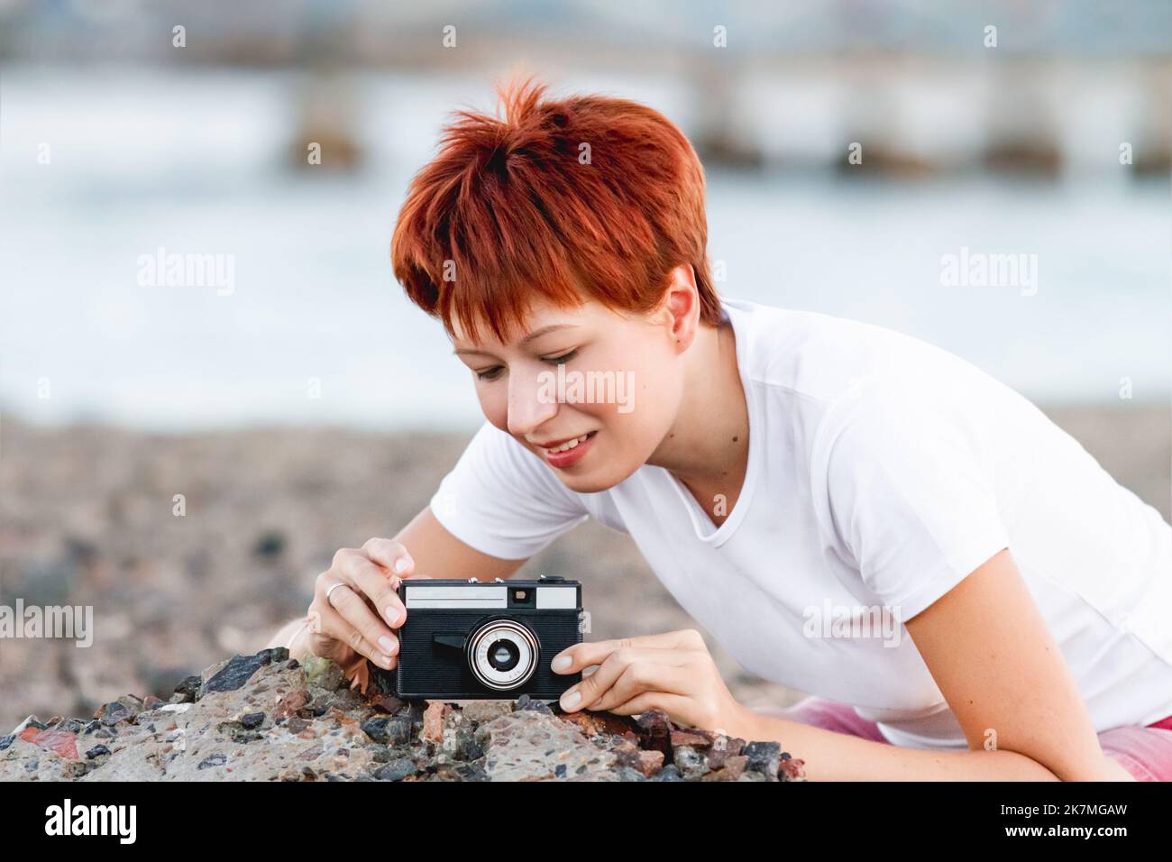 Une femme aux cheveux rouges courts tourne sur un appareil photo de film vintage. La photographie comme passe-temps. Appareil photo rétro avec objectif manuel. Banque D'Images