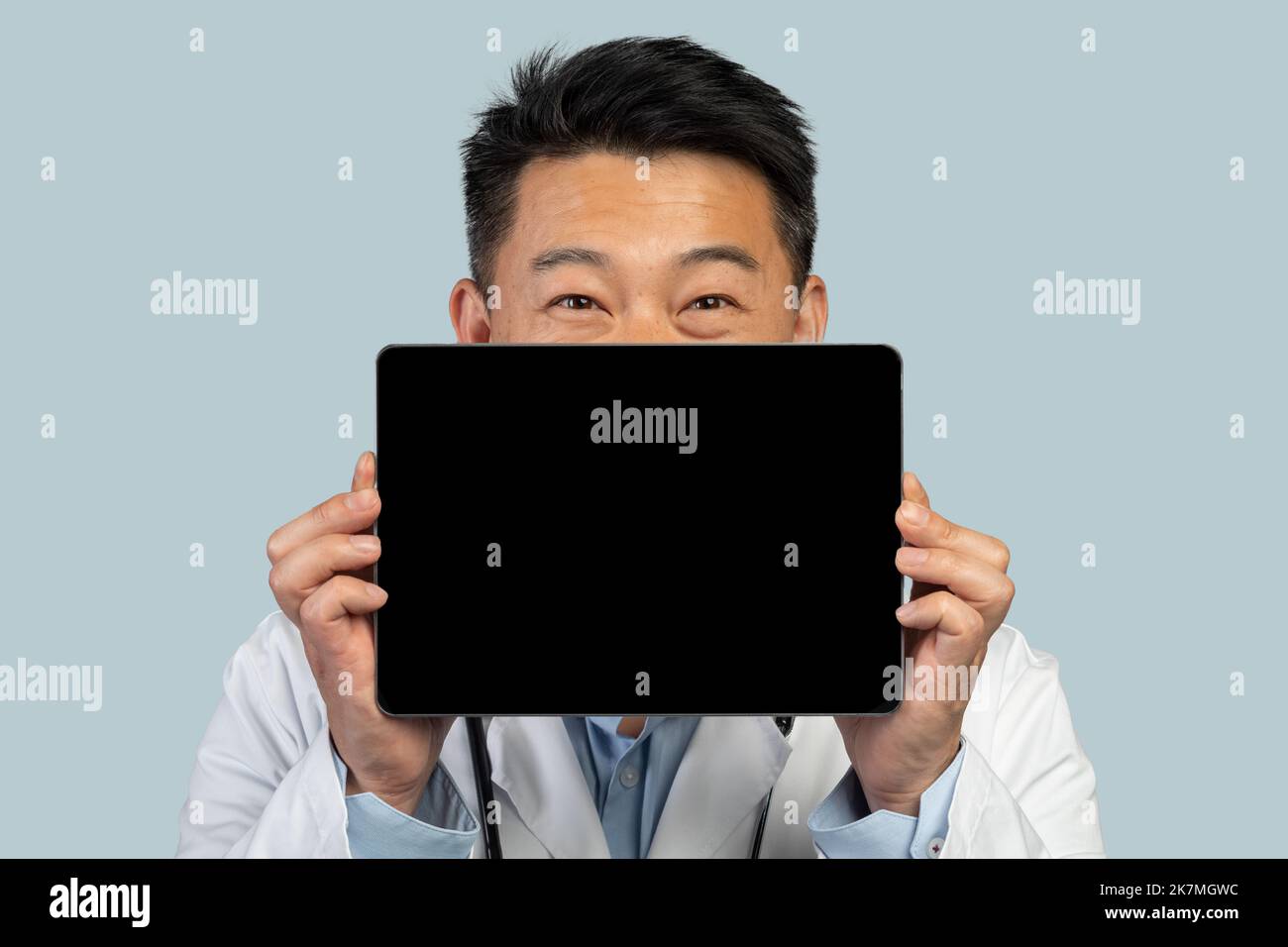 Un homme asiatique d'âge moyen souriant, sous un manteau blanc, montre une tablette avec écran vide Banque D'Images