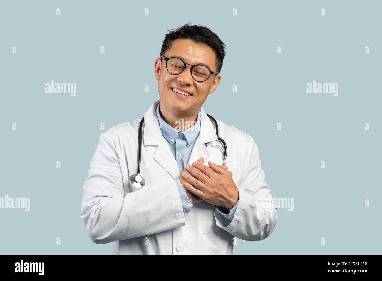 Gaie médecin chinois d'âge moyen en manteau blanc, les lunettes se pressent les mains sur la poitrine Banque D'Images