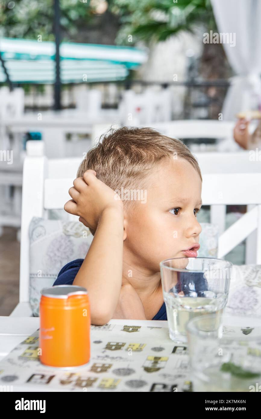 Un garçon d'âge préscolaire est assis dans le restaurant en attendant l'ordre de commencer à manger. Un enfant aux cheveux justes regarde avec une expression ennuyeuse assis à une table en bois Banque D'Images