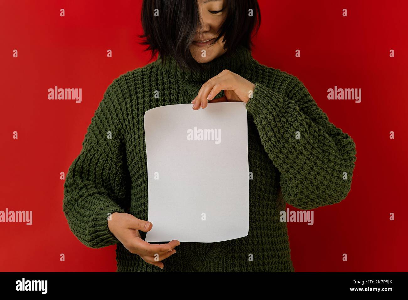 Femme asiatique dans un chandail vert sur un fond rouge tient une feuille de papier vierge dans ses mains, espace de copie. Toile de fond pour la rédaction des objectifs pour l'année, a f Banque D'Images