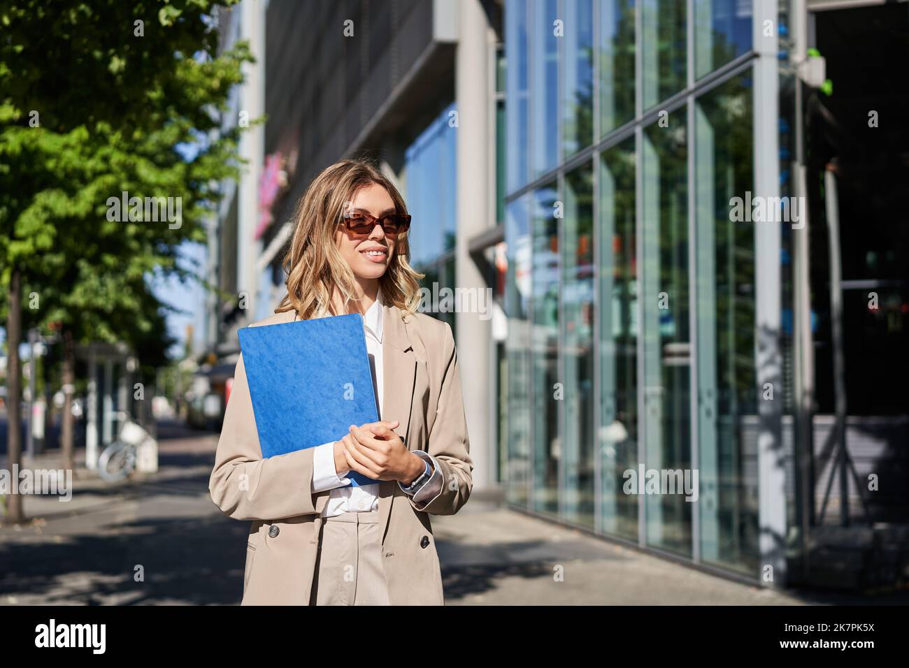 Portrait d'une femme d'affaires en lunettes de soleil et costume beige,  tenant une chemise bleue avec des documents de bureau, allant au travail,  marchant dans la rue le jour ensoleillé Photo Stock -