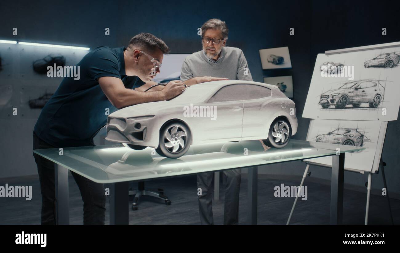 Les ingénieurs de conception automobile senior travaillent sur le modèle prototype de la voiture en regardant les croquis au crayon de la conception. Utilisation d'un outil rotatif pour les retouches finales dans la sculpture du râteau placée sur une table. Banque D'Images