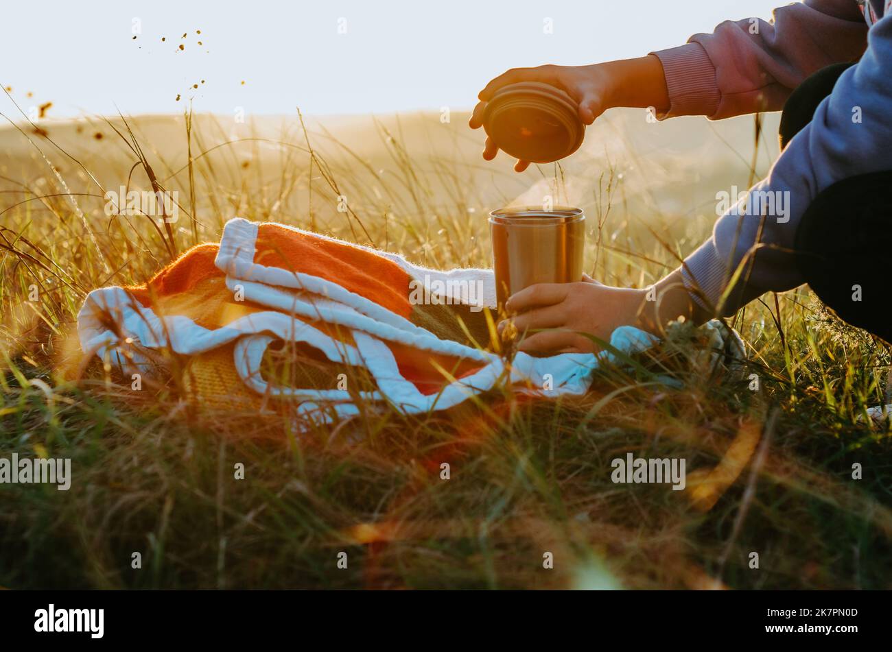 Personne ouvrant le couvercle de la tasse thermo avec thé chaud sur couverture tricotée, sur la colline au coucher du soleil Banque D'Images