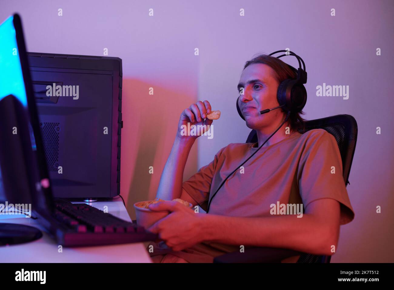 Un jeune homme souriant mangeant des chips de pomme de terre en regardant le flux en direct de blogger préféré sur l'écran d'ordinateur Banque D'Images