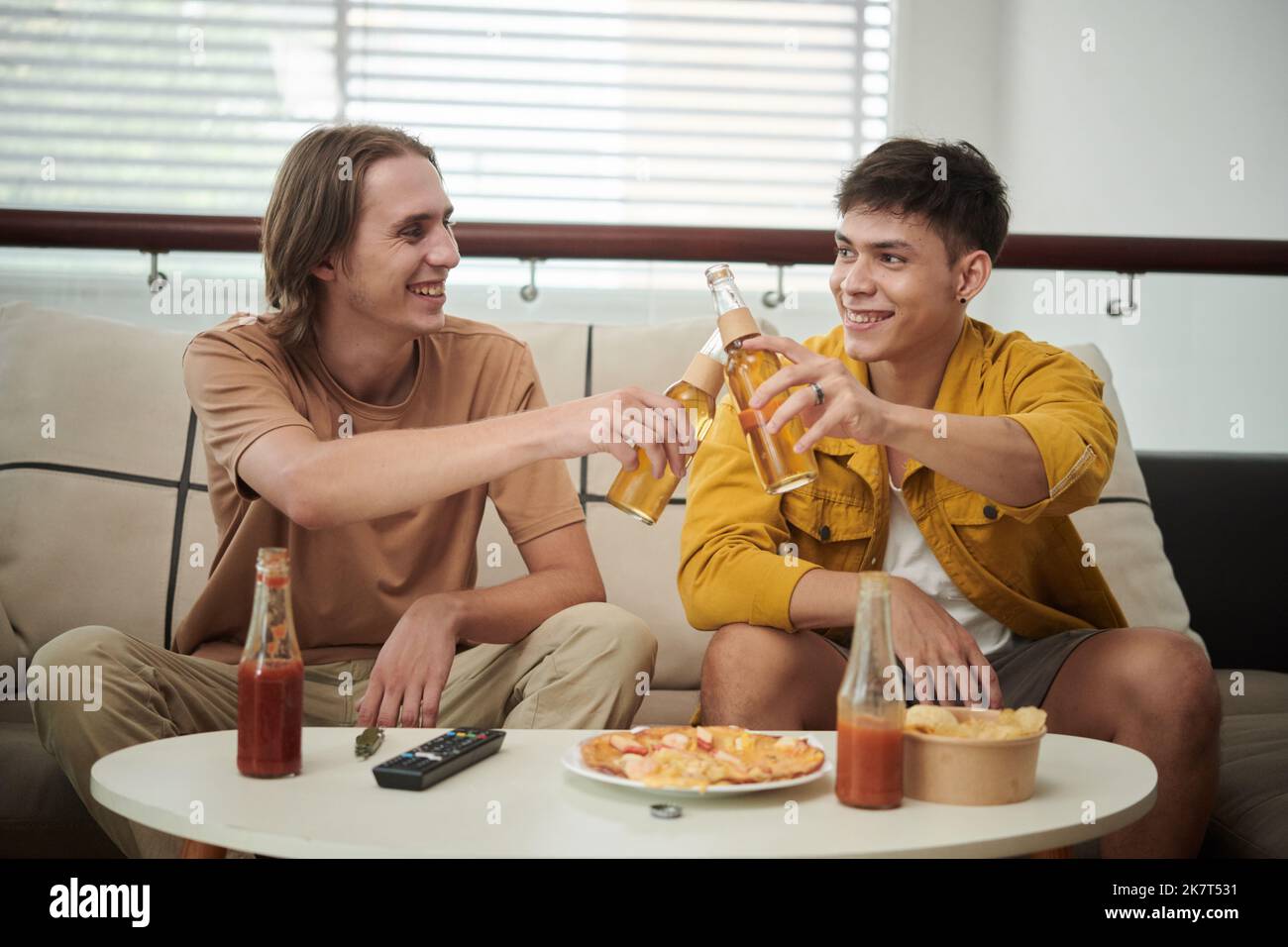 Des amis joyeux se sont empatés des bouteilles de bière sur la table avec de la pizza et des chips de pommes de terre Banque D'Images
