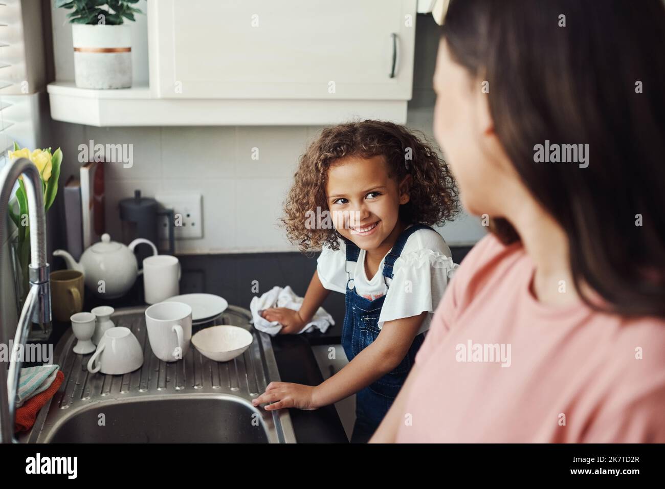 Puis-je jouer maintenant. une adorable jeune fille debout et aidant sa mère avec les plats dans la cuisine à la maison. Banque D'Images