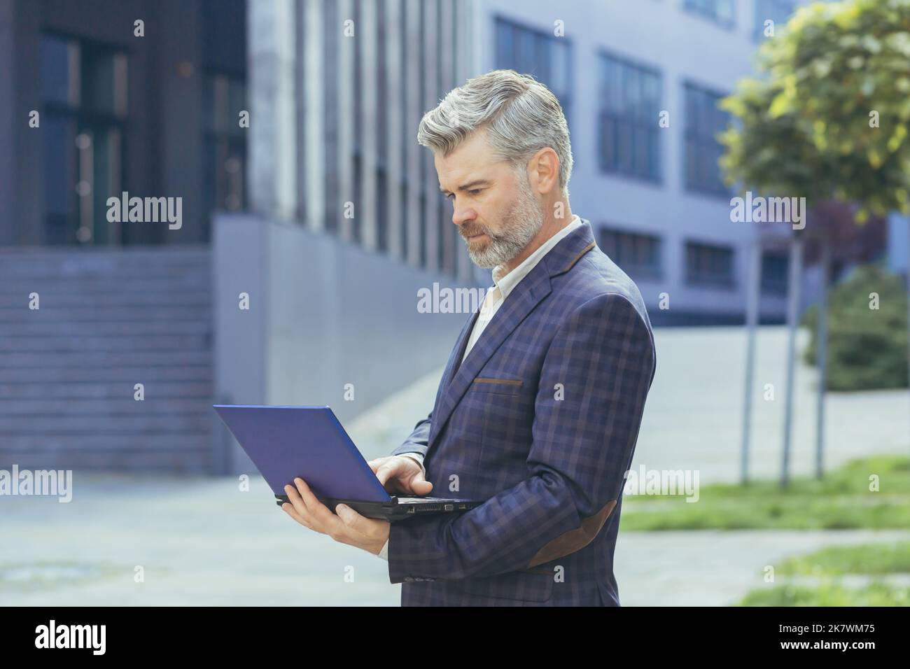 boss gris sérieux et concentré travaillant sur un ordinateur portable en dehors d'un immeuble de bureau moderne, boss mature en costume d'entreprise dactylographiant sur un clavier netbook, homme d'affaires senior réussi. Banque D'Images