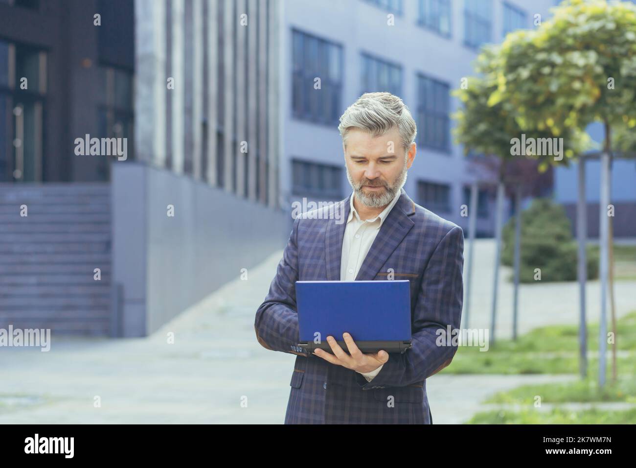 boss gris sérieux et concentré travaillant sur un ordinateur portable en dehors d'un immeuble de bureau moderne, boss mature en costume d'entreprise dactylographiant sur un clavier netbook, homme d'affaires senior réussi. Banque D'Images