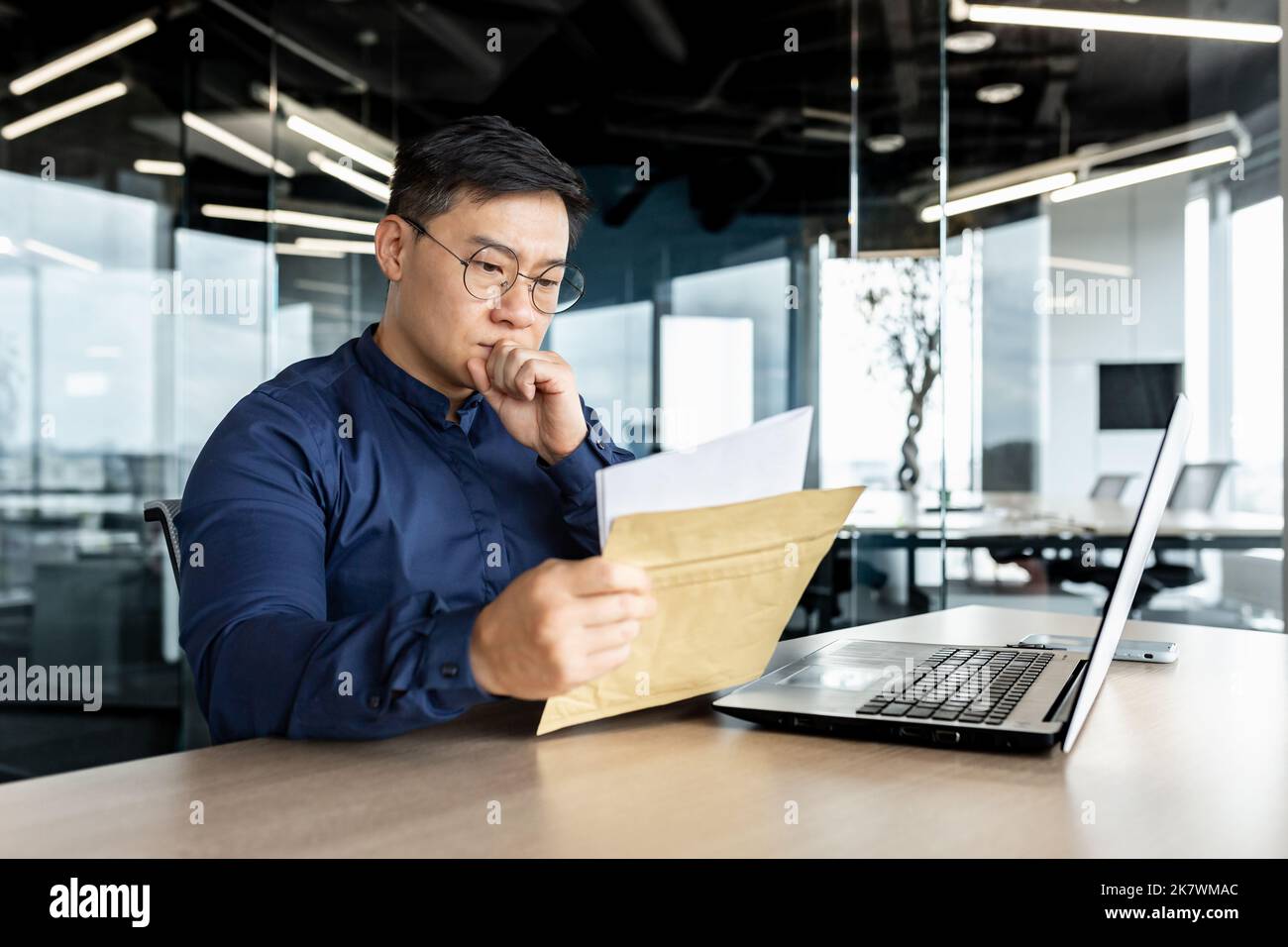 Pensant homme d'affaires asiatique travaillant dans un immeuble de bureaux moderne, homme lisant une lettre reçue de la banque, patron pensant sérieux et concentré travaillant avec un ordinateur portable. Banque D'Images