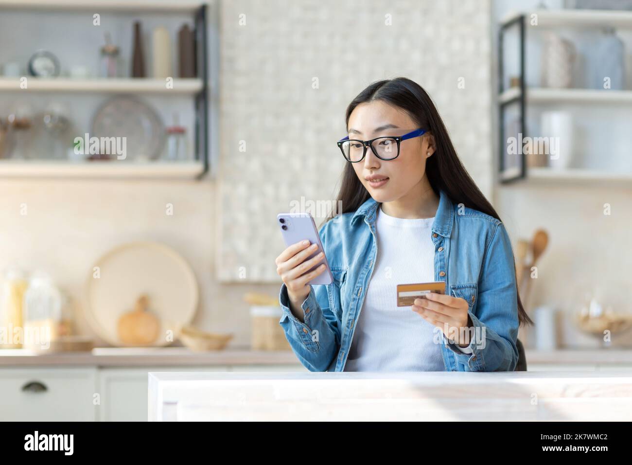 Problèmes financiers. Inquiète jeune femme asiatique tenant le téléphone et la carte de crédit. Assis dans la cuisine à la maison, essayant de résoudre un problème. Banque D'Images