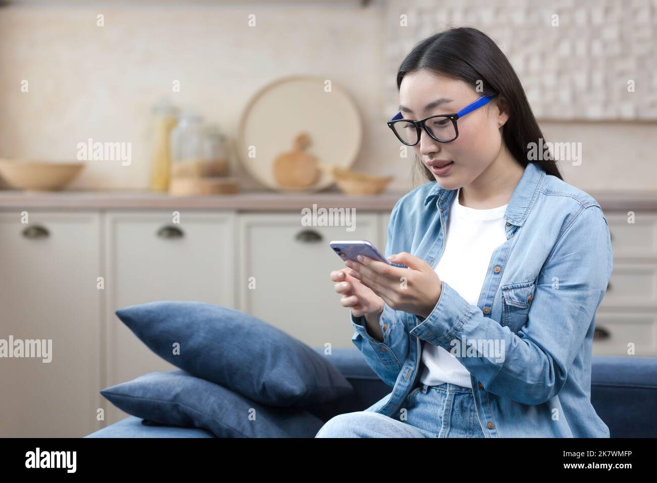 Jeune femme belle à la maison assise dans la cuisine souriante et heureuse, femme asiatique utilisant un smartphone tapant des messages et lisant des nouvelles en ligne. Banque D'Images