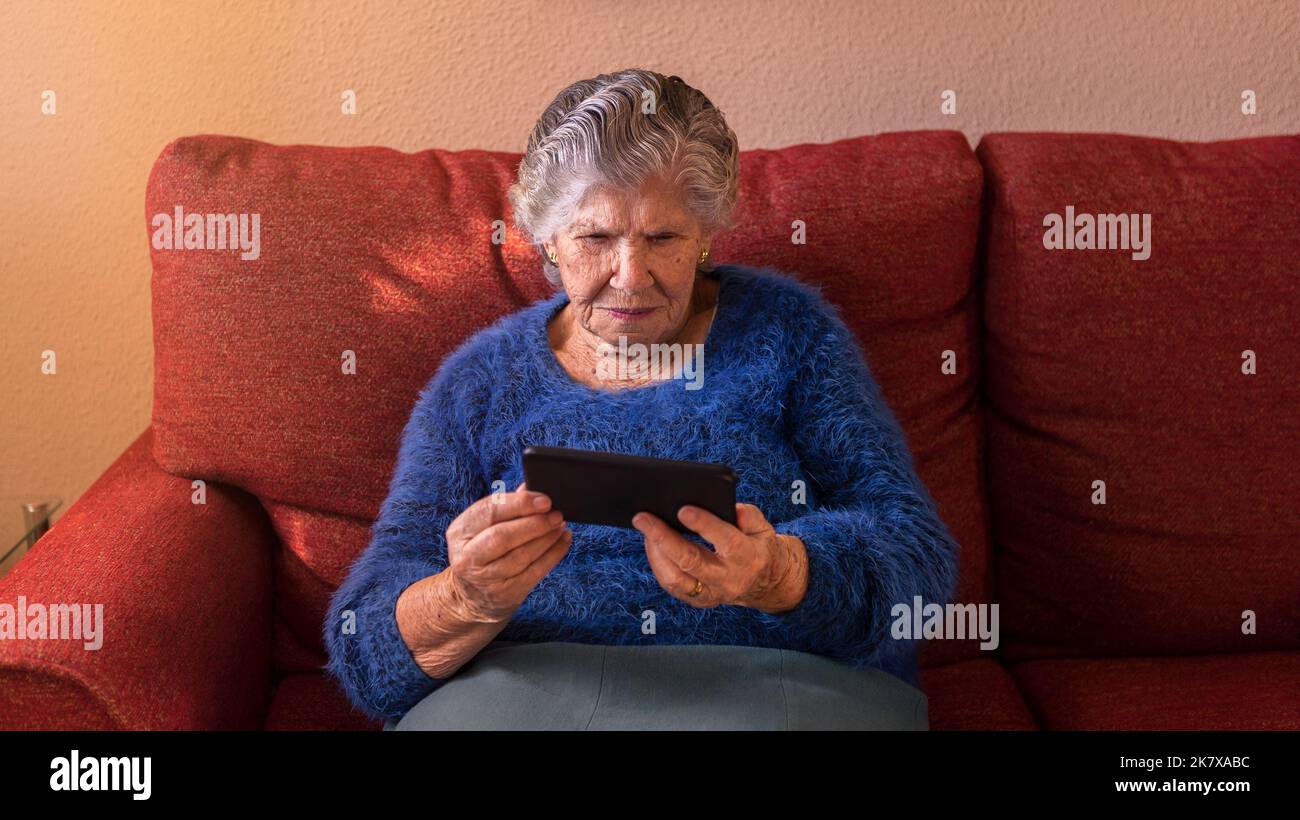 Femme âgée utilisant un téléphone portable tout en étant assise sur un canapé à la maison. Grand-mère tient un smartphone. Ancien grand-mère utilisant un téléphone. Concept de technolog Banque D'Images