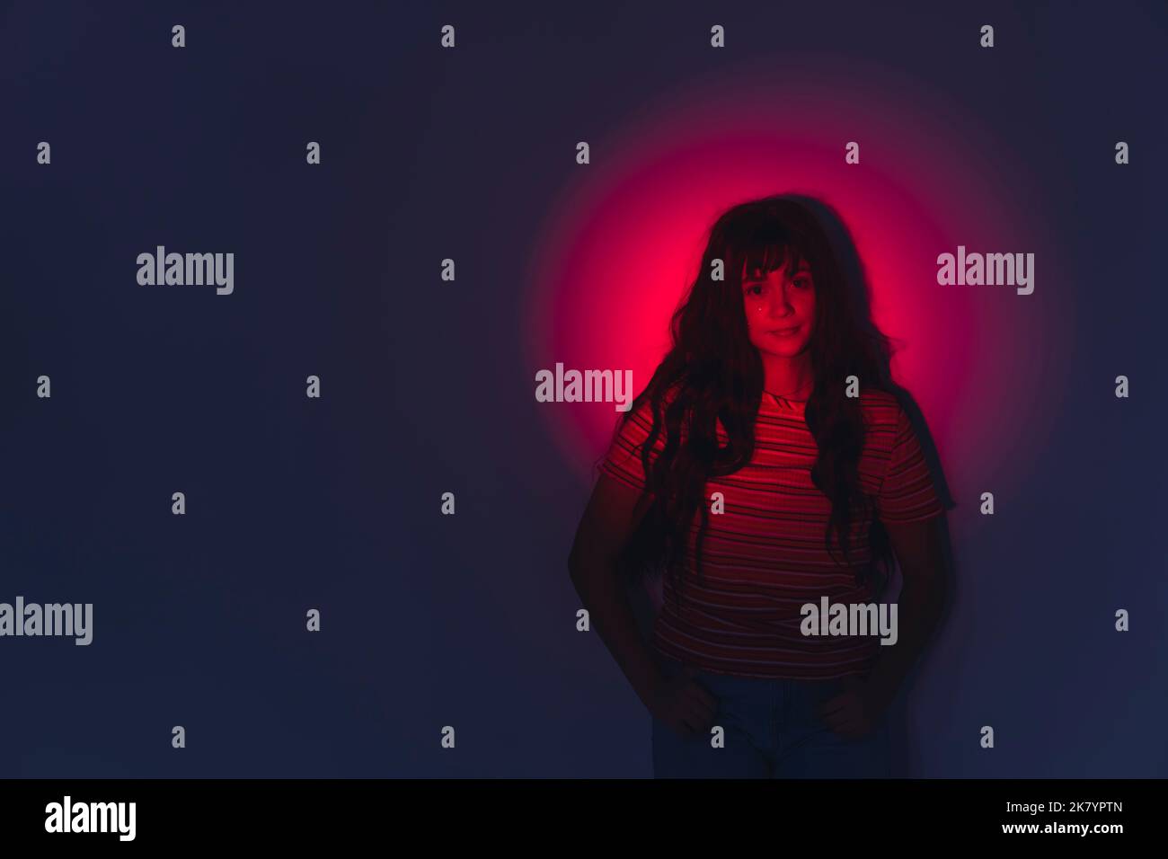 Portrait artistique créatif d'une femme aux cheveux foncés debout devant un mur dans l'obscurité avec un spot rouge sur son visage. Prise de vue horizontale en studio. Photo de haute qualité Banque D'Images