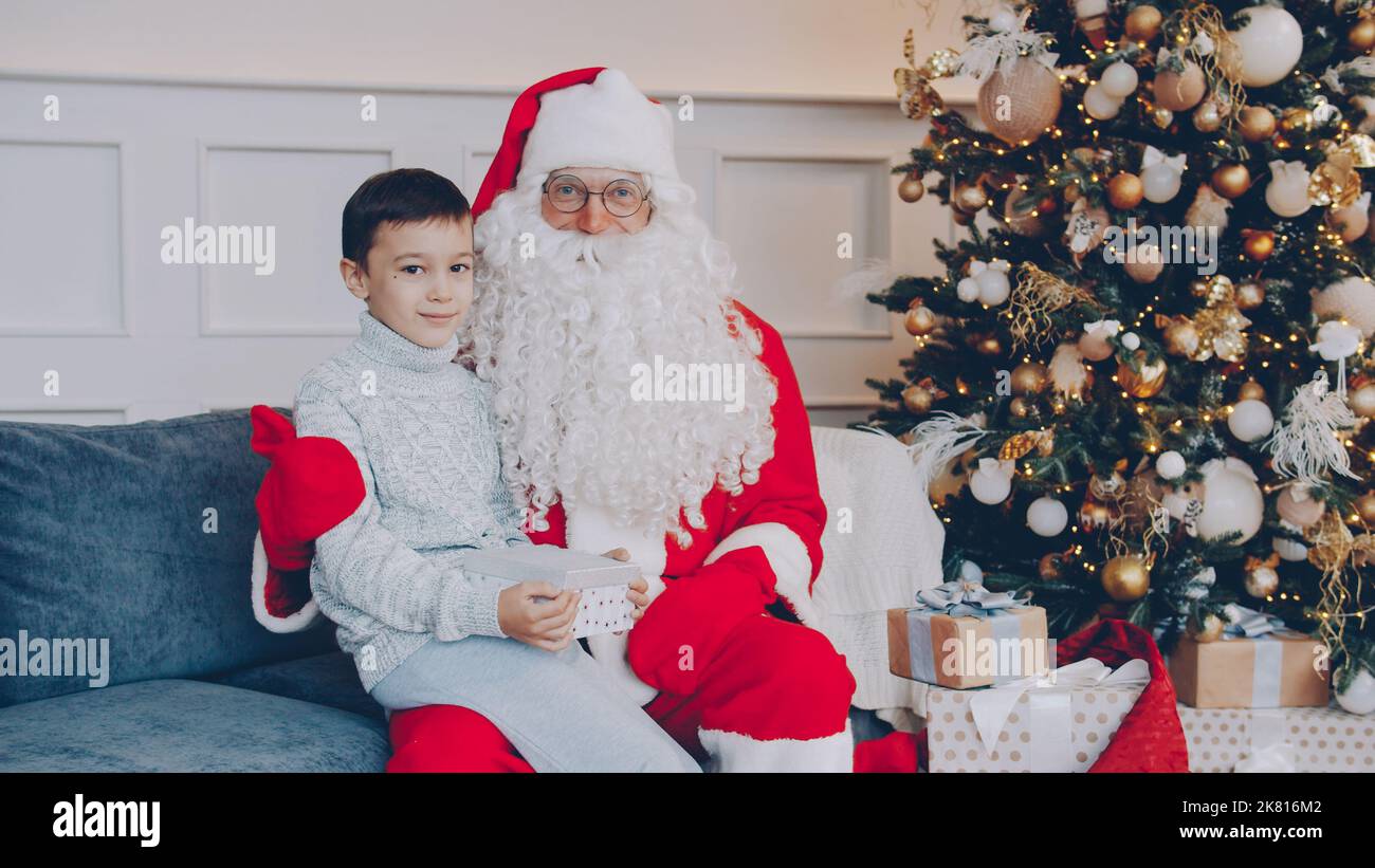 Portrait d'un enfant adorable avec le présent et le Père Noël assis sur un canapé à la maison, regardant l'appareil photo le jour de l'an. Concept de personnes, de célébrations et de cadeaux. Banque D'Images