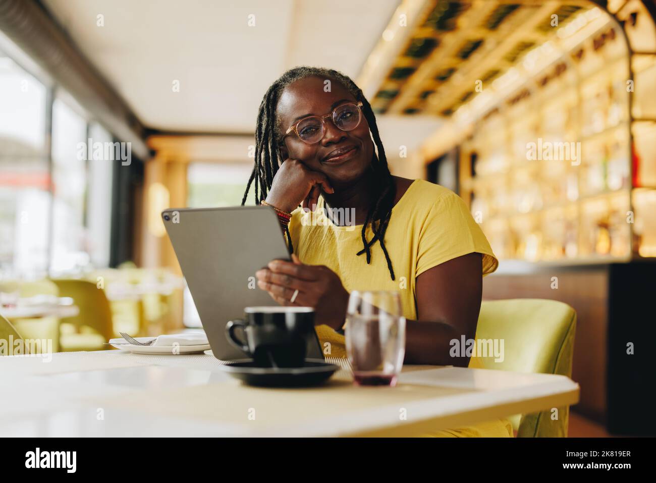 Femme d'affaires mature regardant la caméra tout en tenant une tablette numérique dans un café. Femme indépendante senior faisant un certain travail indépendant en ligne dans un Banque D'Images
