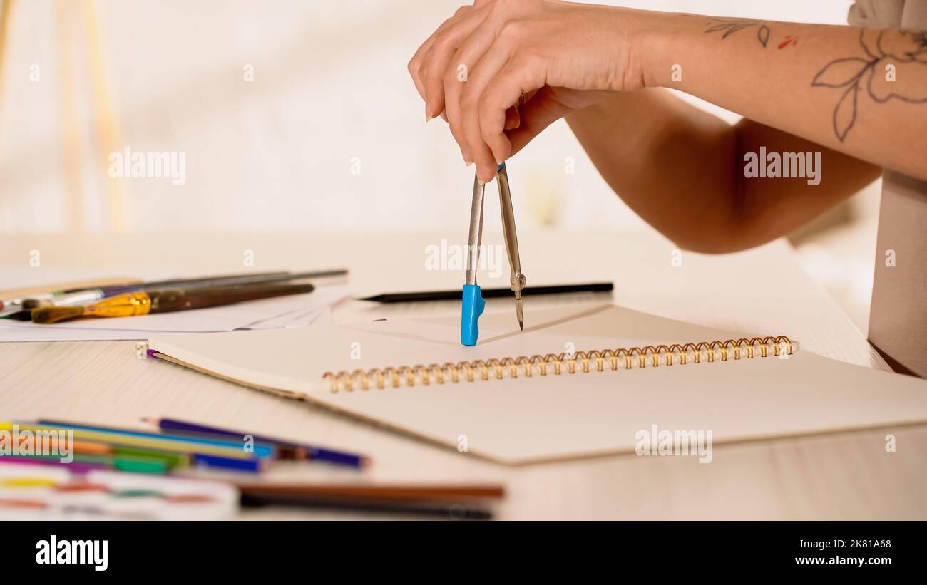 Vue rognée de la femme avec tatouage tenant la boussole de dessin près de l'esquisse à la maison, image de stock Banque D'Images