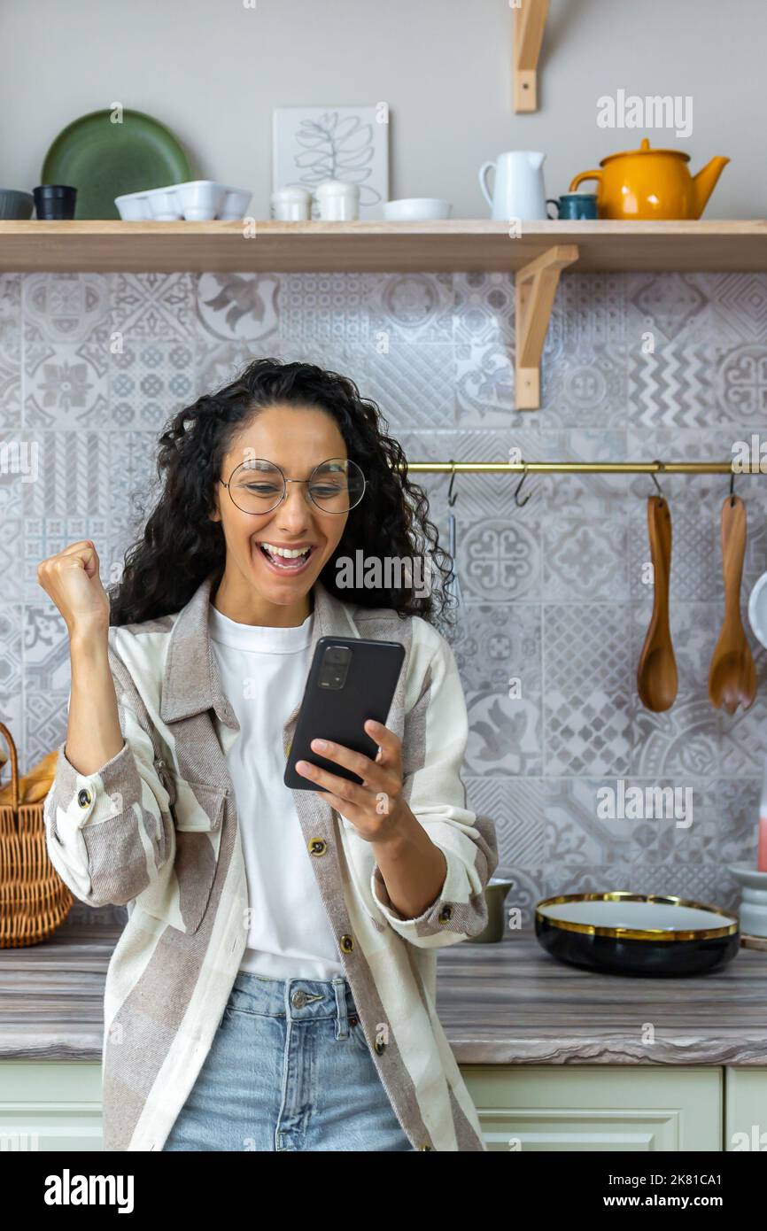 Vertical shot, une femme latino-américaine à la maison dans la cuisine a eu de bonnes nouvelles en ligne, une femme avec des cheveux et des lunettes mauries célébrant la victoire succès en tenant un smartphone. Banque D'Images