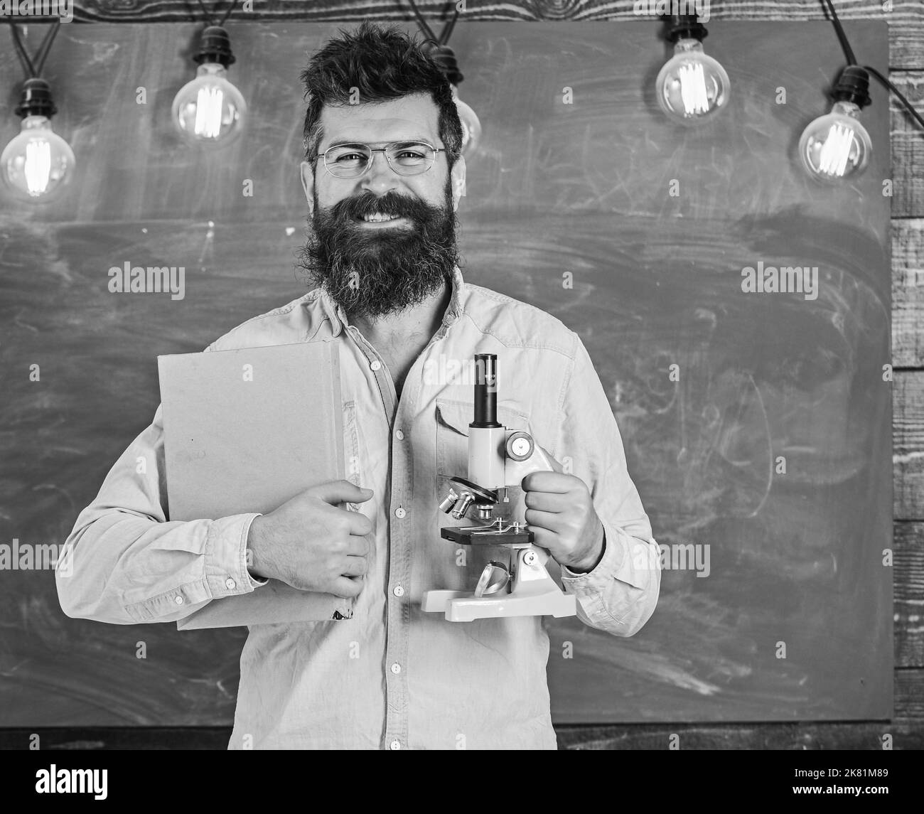 Le professeur de lunettes tient un livre et un microscope. Concept de la science. Homme avec barbe et moustache sur le visage souriant dans la salle de classe. Le scientifique tient livre et Banque D'Images