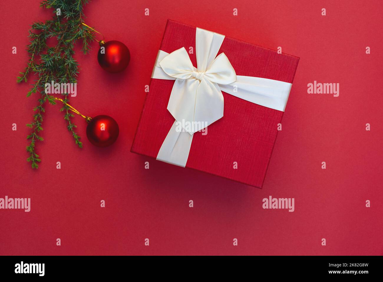 Boîte cadeau avec noeud en ruban et décorations de Noël sur branche d'arbre sur fond rouge. Jouets ronds. Style minimaliste. Copier l'espace. Préparation de cadeaux pour les vacances. Vierge pour la publicité Banque D'Images
