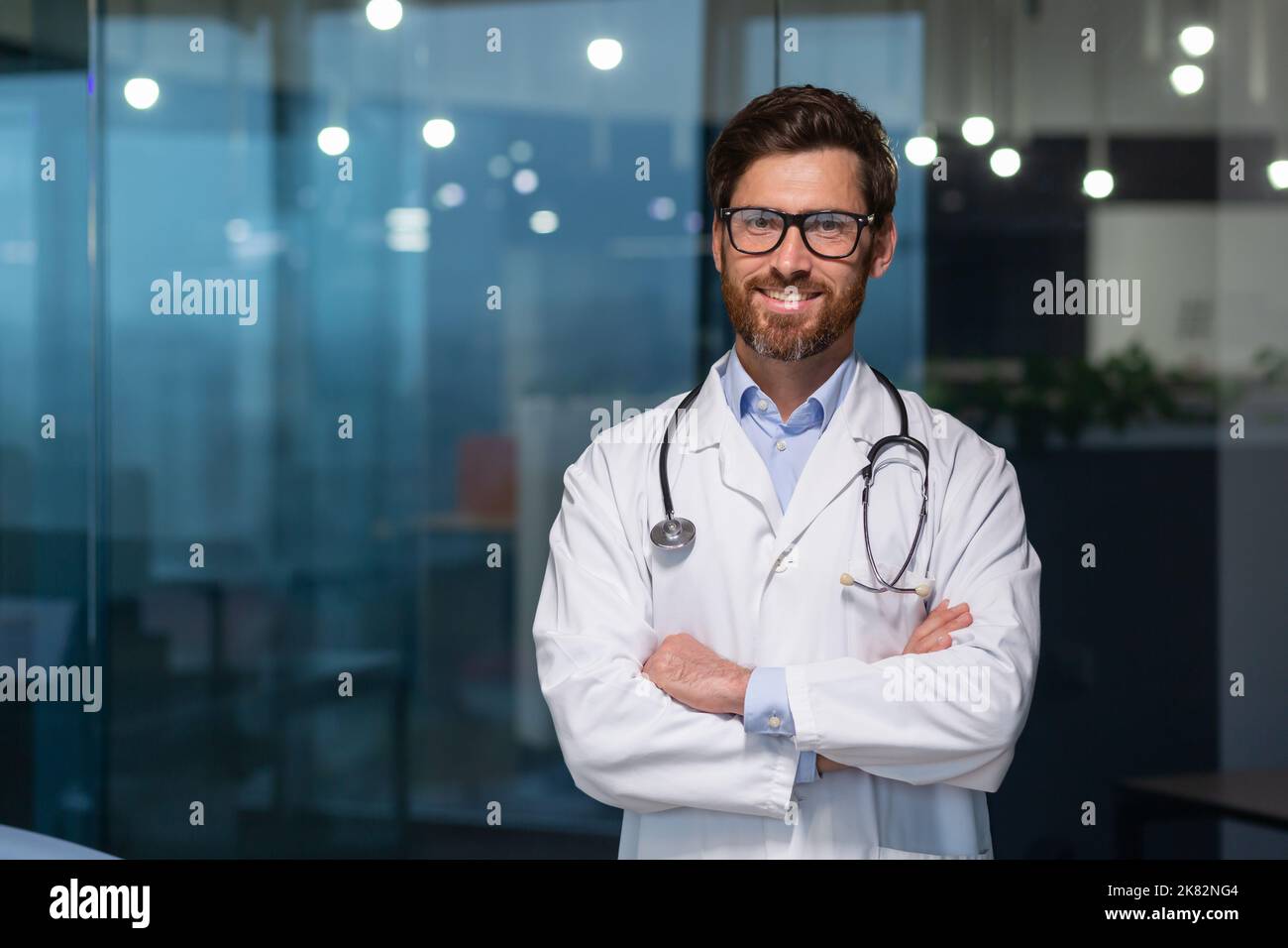 Portrait d'un médecin mature avec barbe, homme en blanc, manteau médical souriant et regardant la caméra avec bras croisés travaillant dans une clinique moderne. Banque D'Images