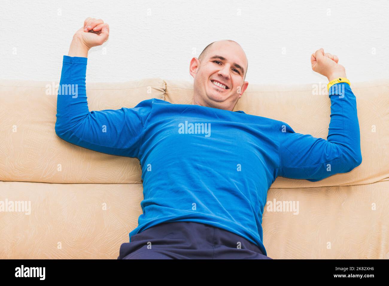Un homme casien bald vêtu d'un t-shirt bleu à manches longues s'étire sur un canapé. Banque D'Images