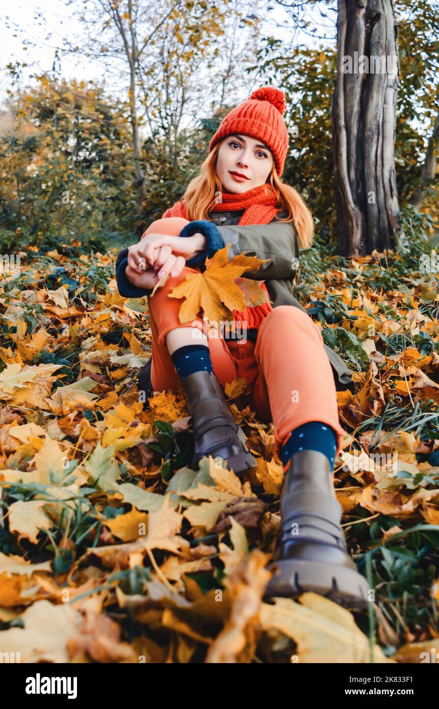 Jeune femme lumineuse et mignonne en tricot orange chaud chapeau et foulard sur fond de feuilles jaunes d'automne. Vue à angle bas Banque D'Images